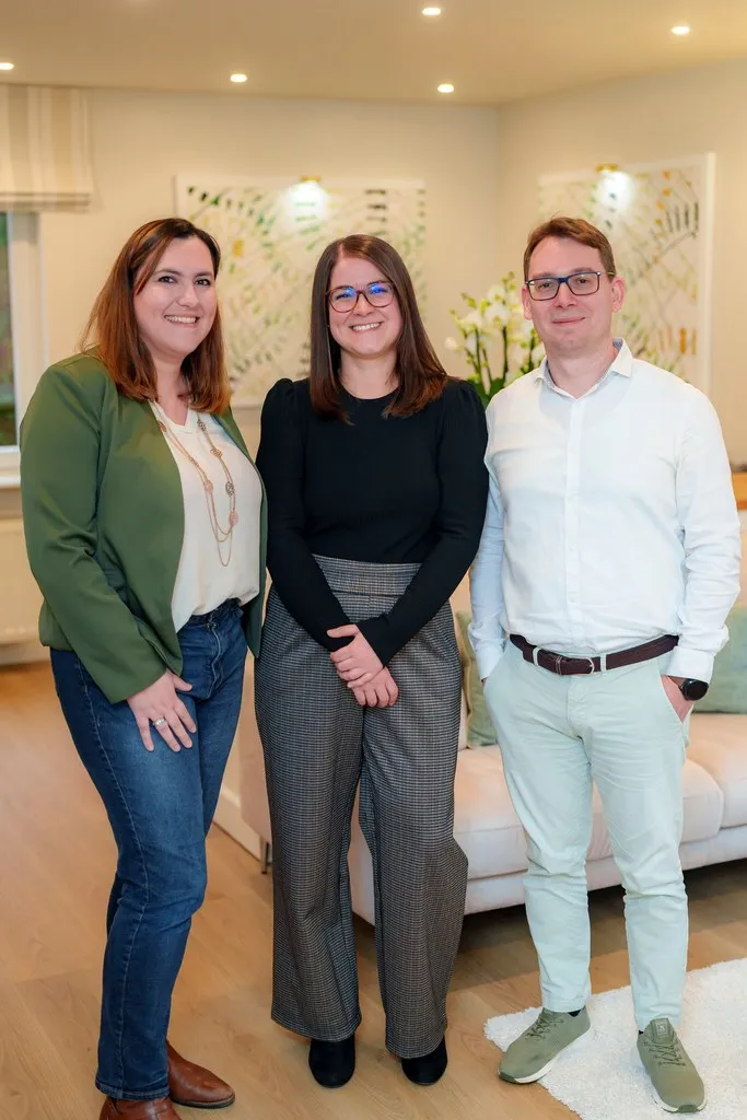 Trois adultes se tiennent à l'intérieur, souriant à l'appareil photo - peut-être en train de célébrer leur succès avec une application organisation famille pour une gestion familiale intelligente. Chacun affiche son style personnel dans des tenues colorées et décontractées.