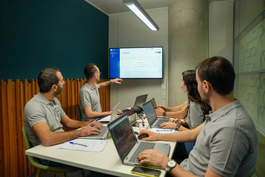 Cinq personnes en chemise grise sont assises autour d'une table de conférence avec des ordinateurs portables, regardant un homme pointer un grand écran affichant une application organisation famille pour la centralisation de documents personnels dans une salle de réunion moderne.