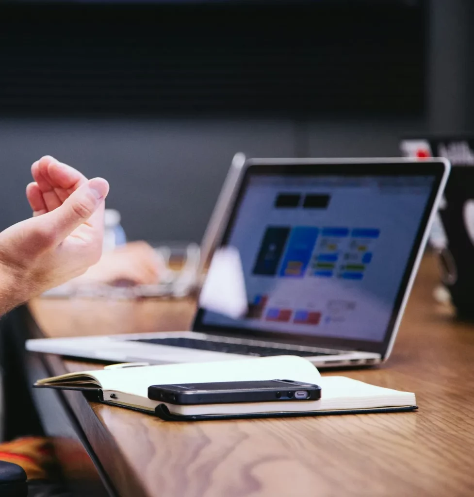 Une personne fait des gestes de la main alors qu'elle est assise à une table en bois avec un ordinateur portable ouvert affichant un écran coloré, un carnet de notes et un smartphone - des outils parfaits pour la gestion familiale intelligente et la centralisation des documents personnels.
