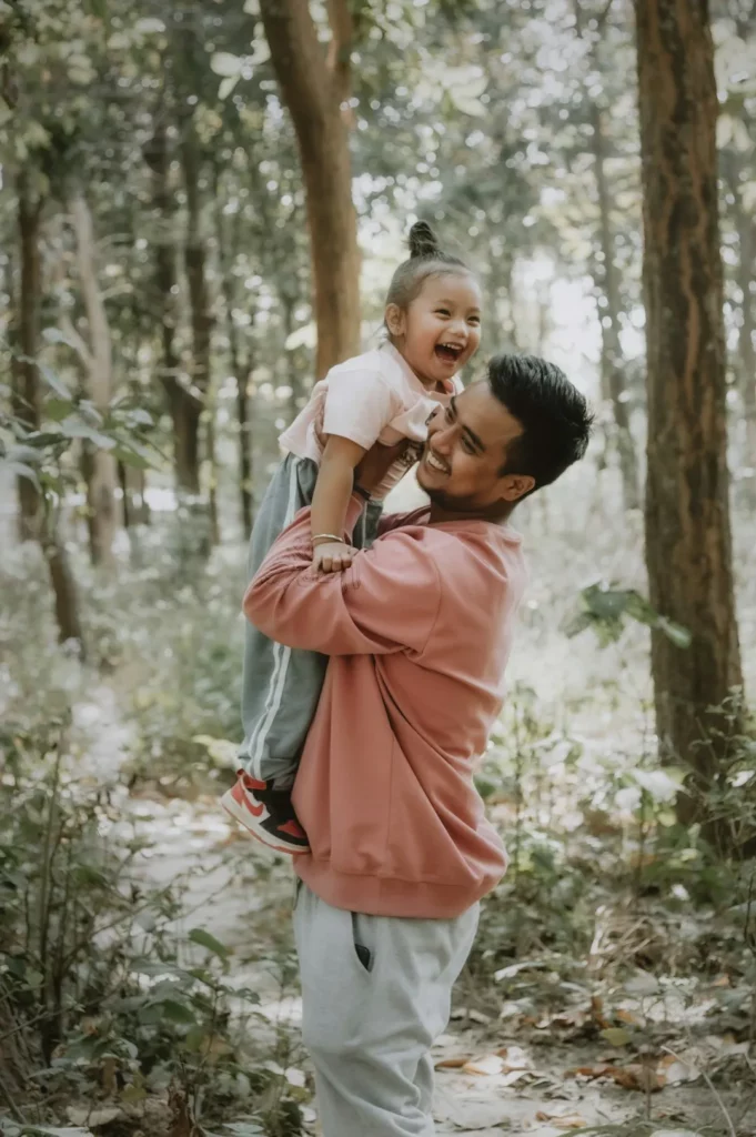 Un homme vêtu d'un sweat-shirt rose soulève une jeune fille souriante en tenue décontractée dans une forêt ensoleillée. Tous deux semblent joyeux, à l'image des familles qui bénéficient de la gestion familiale intelligente pour simplifier la vie quotidienne.