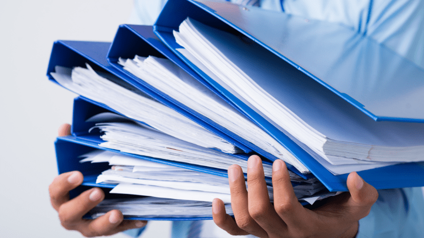 Une personne tenant plusieurs classeurs bleus épais remplis de piles de papier, ce qui suggère une charge de travail importante ou une documentation considérable - soulignant la nécessité de centraliser les documents personnels.