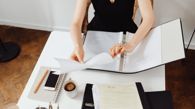 Une personne est assise à un bureau, feuilletant des documents dans un classeur, se demandant peut-être combien de temps doit-on garder les factures d'eau. Sur le bureau se trouvent un cahier avec un crayon, un smartphone, une tasse de café et plusieurs dossiers contenant des documents.