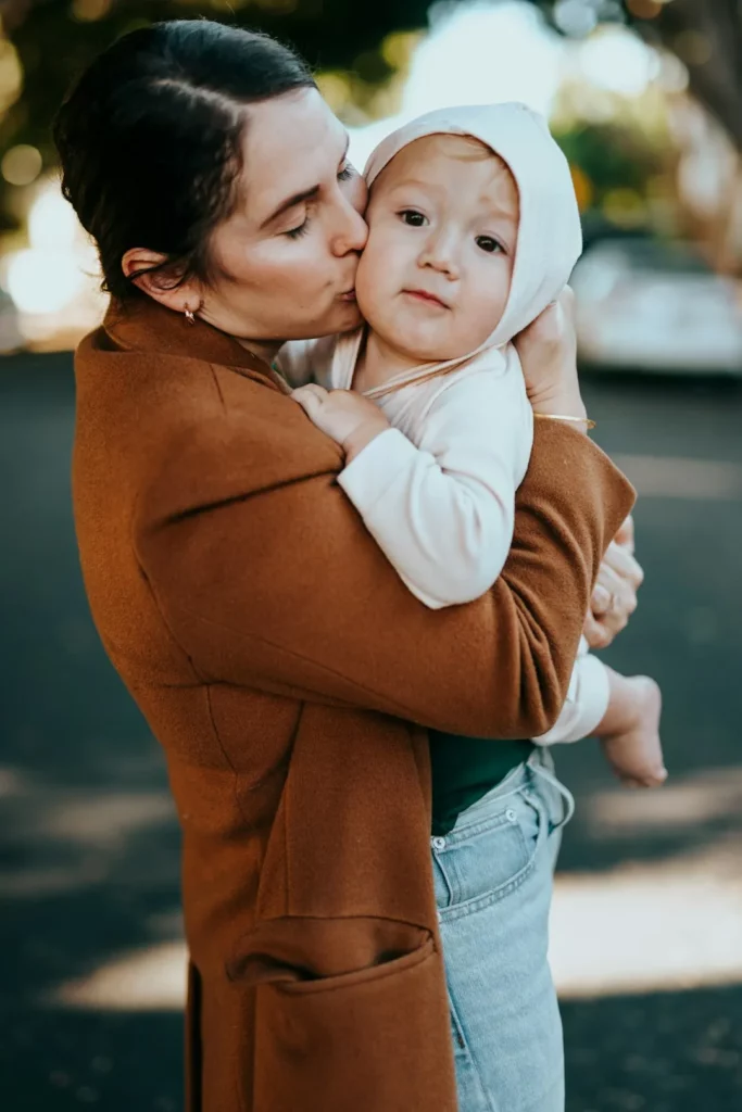 Une femme vêtue d'un manteau marron embrasse affectueusement un bébé sur la joue tout en le tenant dans ses bras. Le bébé, vêtu d'une capuche, regarde calmement l'appareil photo alors qu'ils profitent d'une promenade en plein air - parfait pour les familles utilisant un calendrier familial partagé.
