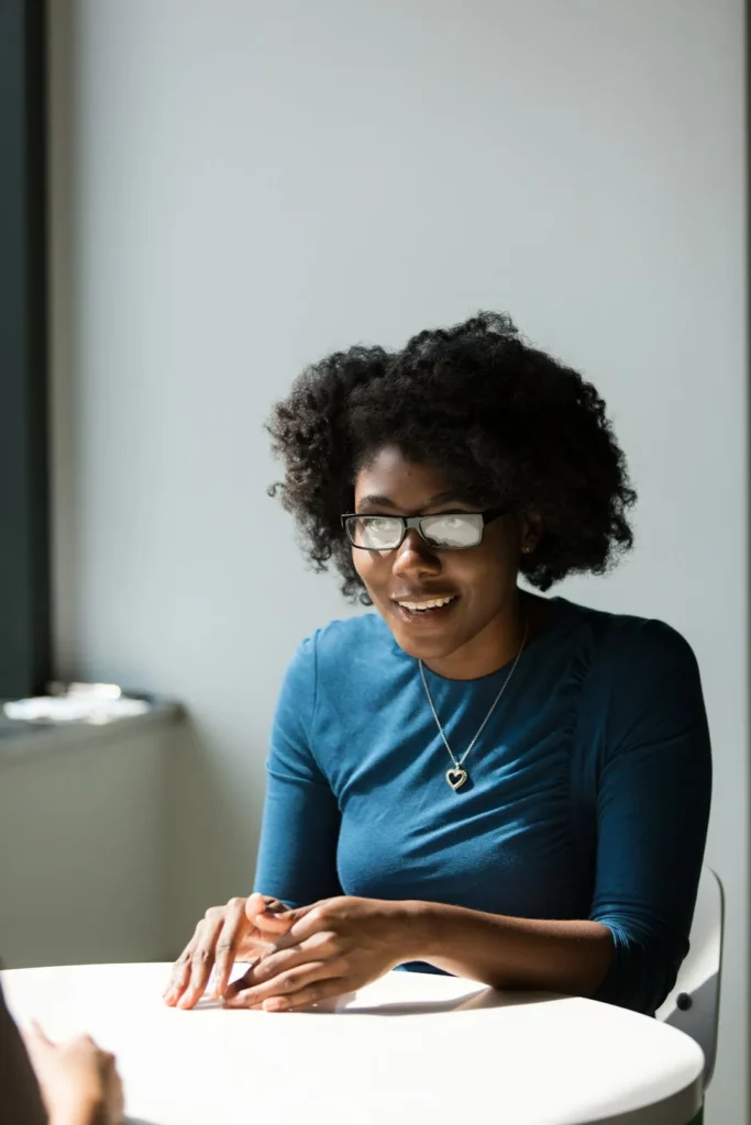 Une femme aux cheveux bouclés et aux lunettes, portant un haut bleu et un collier en forme de cœur, est assise à une table, souriante et semblant engagée dans une conversation sur la gestion tâches et patrimoine familial sur une plateforme organisation familiale.