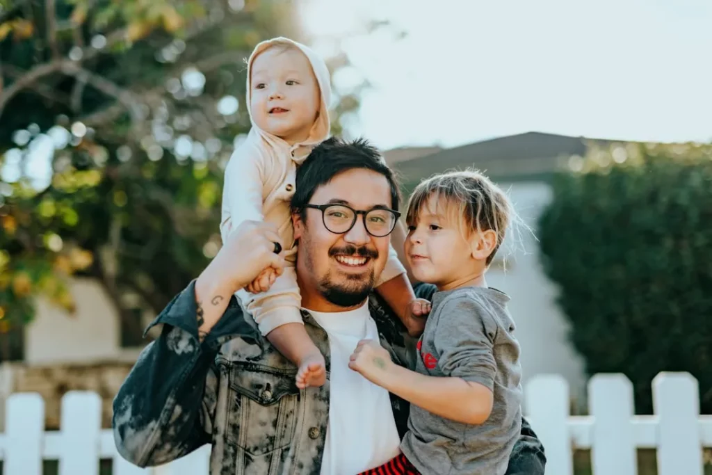 Un homme souriant portant des lunettes tient un enfant en bas âge sur son épaule et serre un enfant plus âgé dans ses bras, le tout en plein air devant une clôture blanche et de la verdure, profitant de la lumière du soleil - tout comme une plateforme d'organisation familiale rassemble tout le monde.
