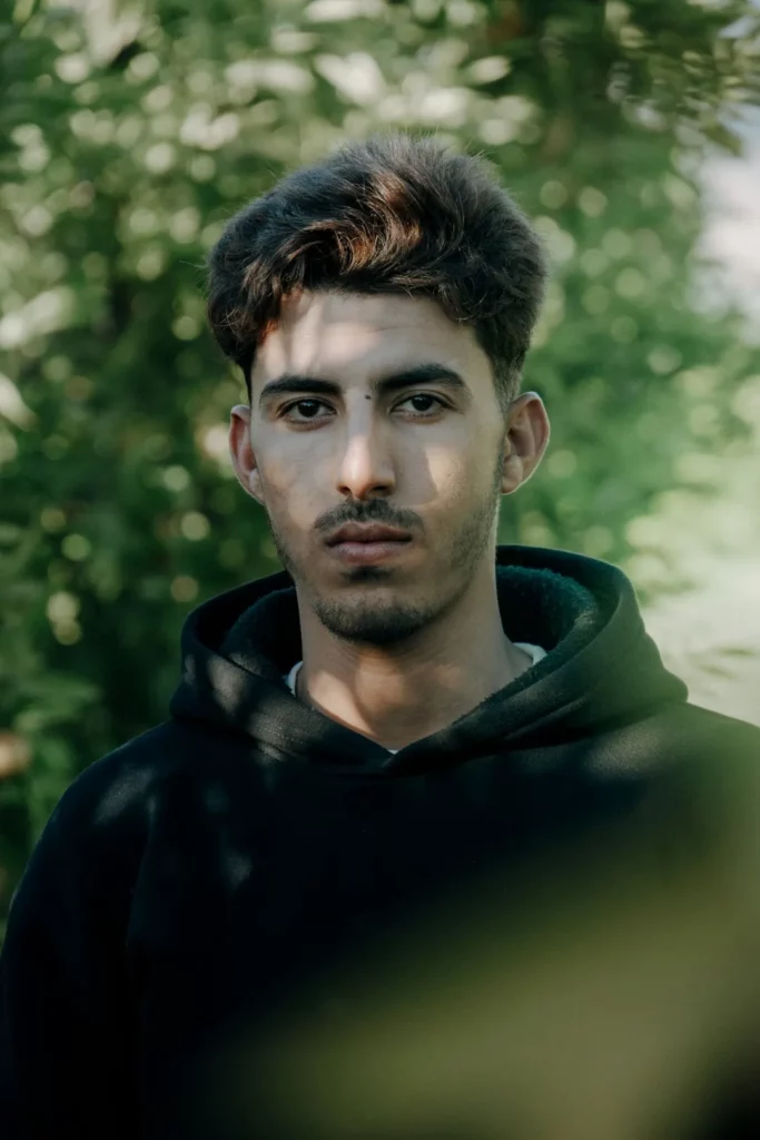 Un jeune homme aux cheveux bruns courts et à la barbe taillée porte un sweat à capuche noir. Il se tient à l'extérieur, devant un feuillage vert flou, et regarde directement l'appareil photo avec une expression neutre, incarnant la mise au point nécessaire à la gestion familiale intelligente.