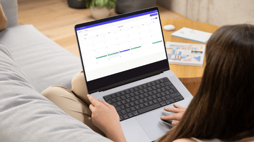 Une personne est assise sur un canapé et utilise un ordinateur portable pour consulter un calendrier partagé permettant d'organiser des événements. Un livre et un stylo reposent sur une table en bois à proximité, mettant en valeur cette solution simple pour créer et gérer des emplois du temps partagés.