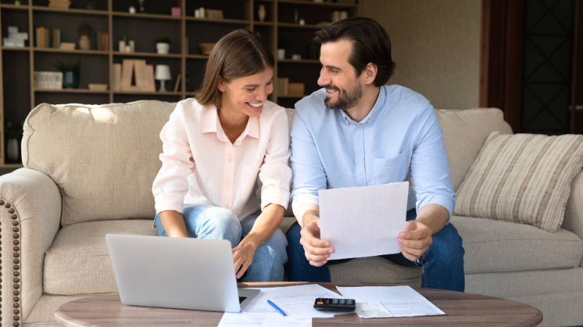Un couple souriant est assis sur un canapé à la maison, examinant des documents ensemble, pratiquant l'organisation de la vie familiale. Un ordinateur portable, une calculatrice et des documents sont posés sur la table basse devant eux. Des étagères contenant des objets de décoration ornent l'arrière-plan.