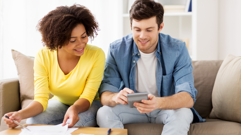 Un couple souriant est assis sur un canapé et examine des documents ensemble ; la femme tient des papiers et un stylo, tandis que l'homme utilise une calculatrice. Ils semblent travailler sur leurs finances ou préparer un Brouillon auto à la maison.