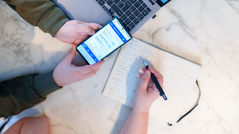 Deux personnes travaillent ensemble à une table ; l'une utilise un smartphone pour consulter une application d'organisation familiale, tandis que l'autre prend des notes dans un carnet. Un ordinateur portable est également visible à proximité.
