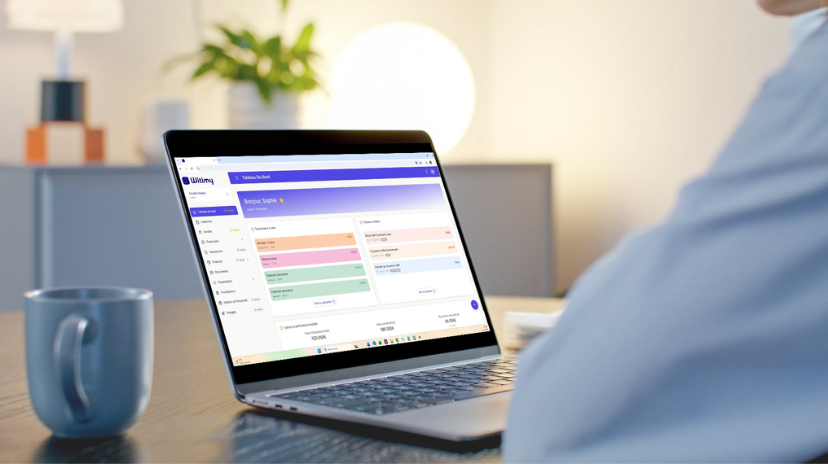 Une personne utilisant un ordinateur portable à un bureau, regardant un tableau de bord coloré sur l'écran - parfait pour la gestion du temps. Une tasse bleue est posée sur le bureau, et l'arrière-plan montre une plante et une lampe rougeoyante dans une pièce doucement éclairée.
